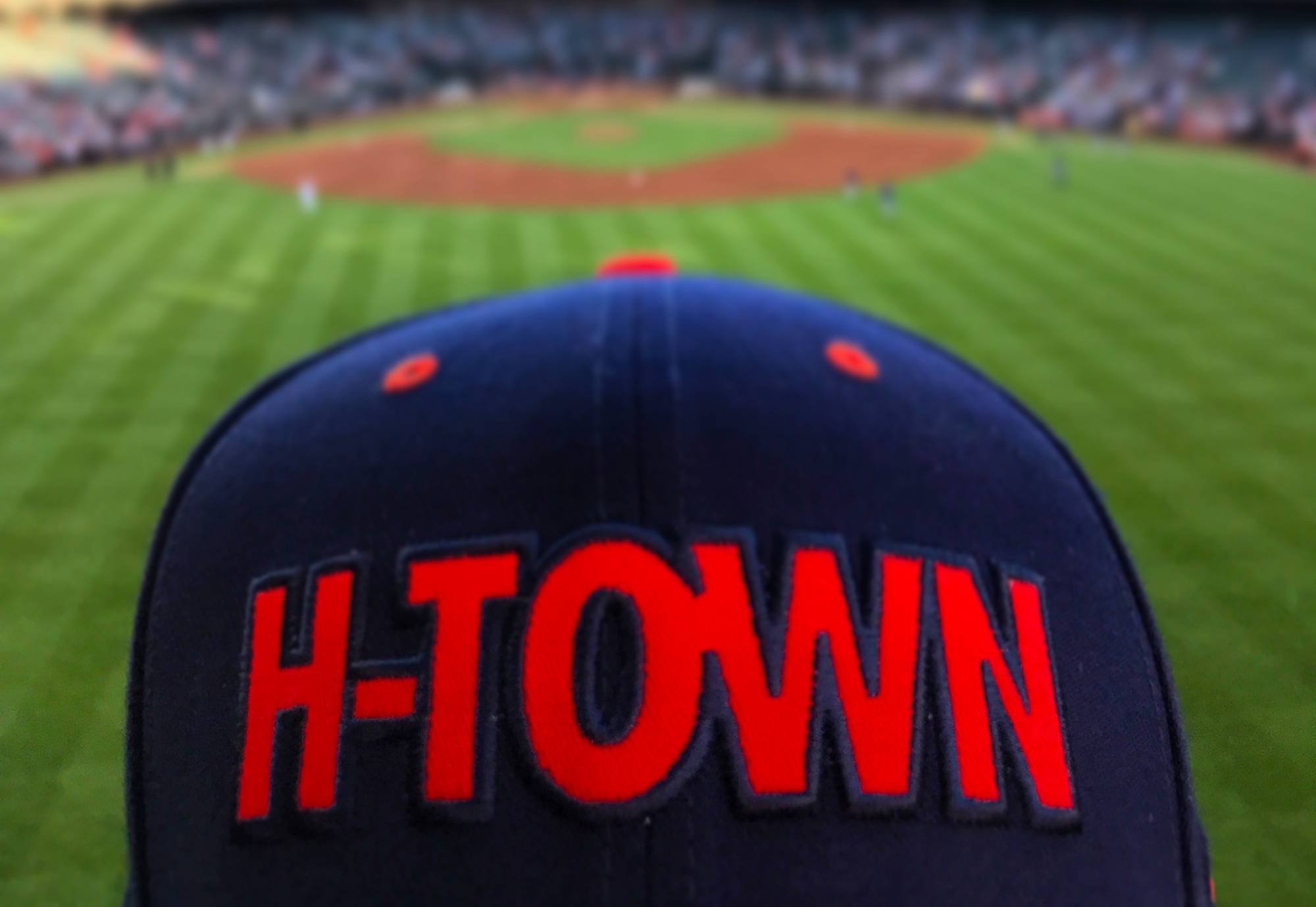 A blue hat with orange block letters reading "H-TOWN" sits in center field of a baseball stadium.